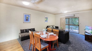A dining area with a round table, chairs, and a sofa. There are two framed pictures on the walls, a ceiling fan, and an air conditioning unit.