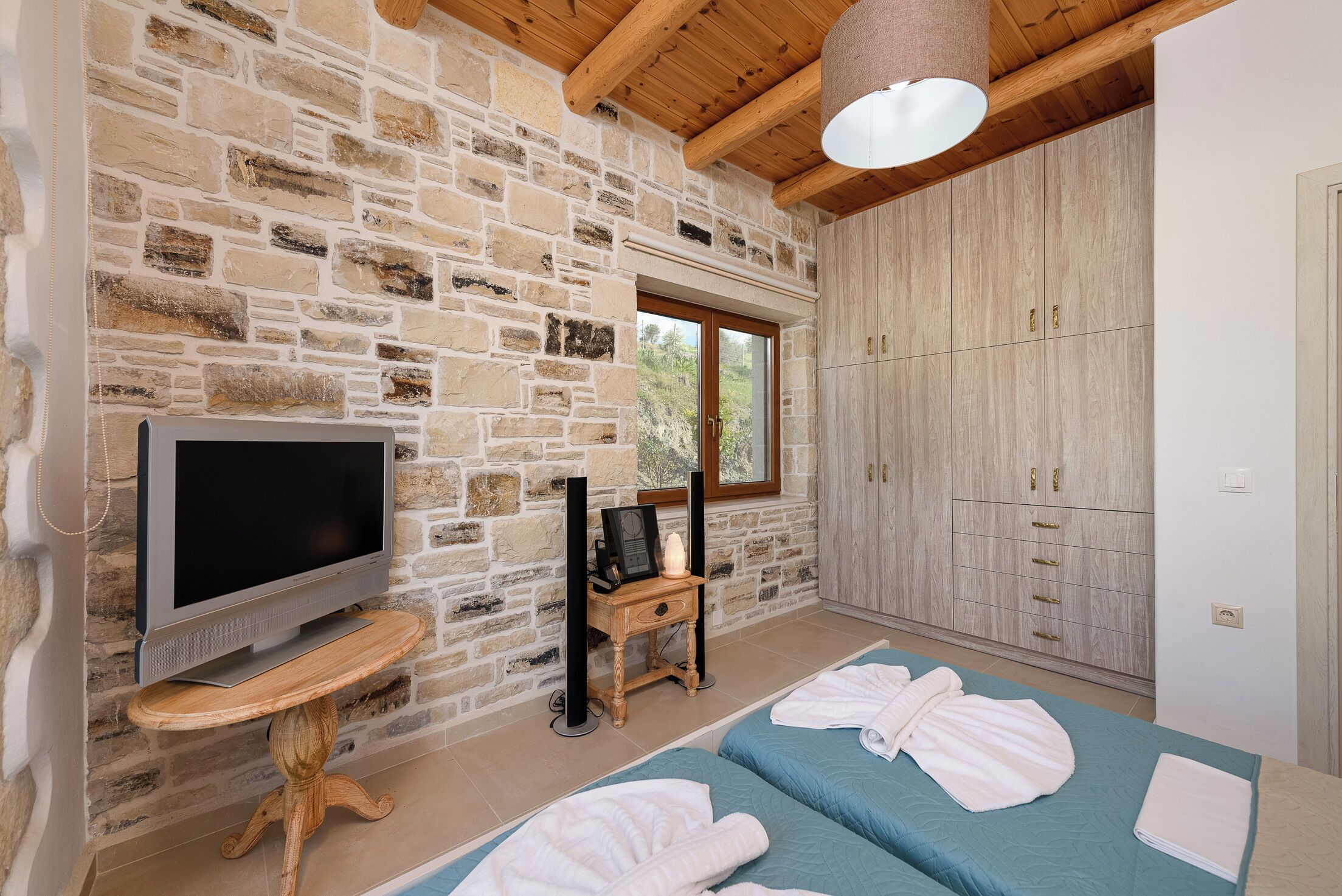 A room with a stone wall, a wooden ceiling, a television on a wooden stand, a wardrobe, and two beds with towels.