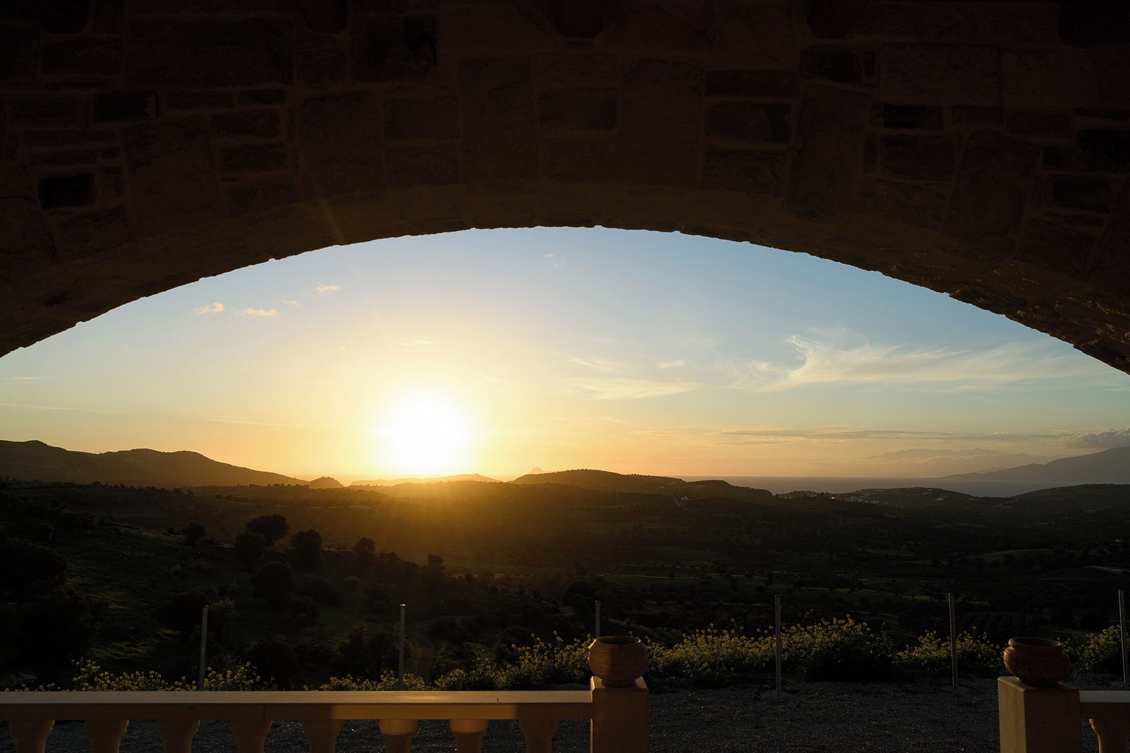 Ein Sonnenuntergangsblick durch eine Bogenkonstruktion aus Stein auf eine hügelige Landschaft.