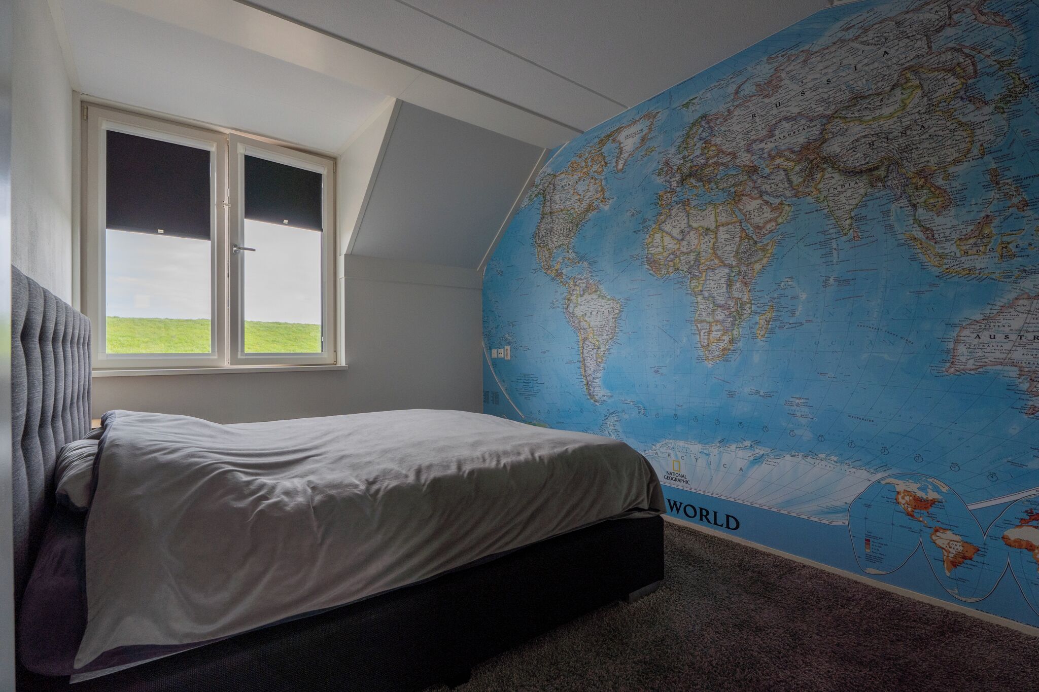 A bedroom with a large world map on the wall, a bed, and a window with a view of the sky.