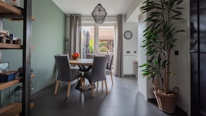 Un espace repas comprenant une table, des chaises, un luminaire suspendu, une plante en pot et une vue sur l’extérieur, visible à travers une porte.