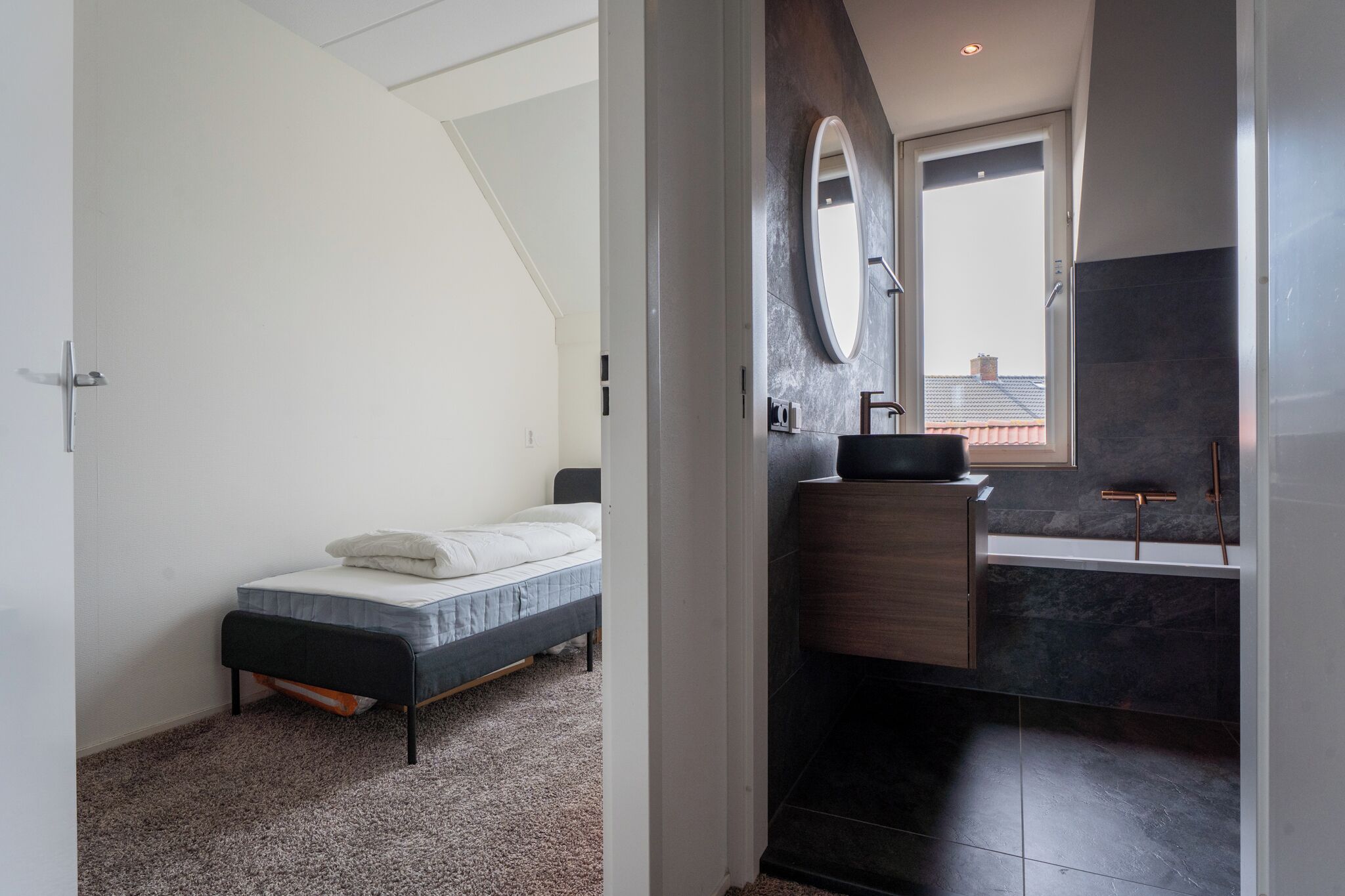 A bedroom with a bed, a bathroom with a sink and mirror, and a view of the outside.