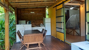 A wooden deck with a table and chairs, a kitchen area with appliances, and a sliding glass door leading to a covered porch. - Charming Bungalow (Le Vauclin)