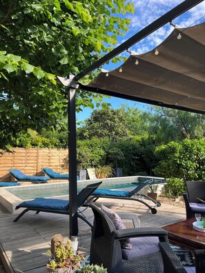 Une terrasse extérieure avec des chaises longues, une piscine et une pergola illuminée de guirlandes lumineuses.
