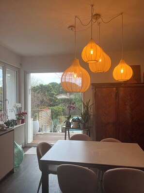 Un coin repas avec une table et des chaises, une vue sur un jardin à travers une porte vitrée et des lampes suspendues.