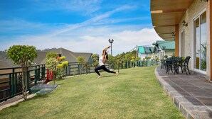 A person performing a yoga pose on a grassy lawn with a building and a fence in the background. - Treetop by Eight Continents Royalpine HP (Kasauli)
