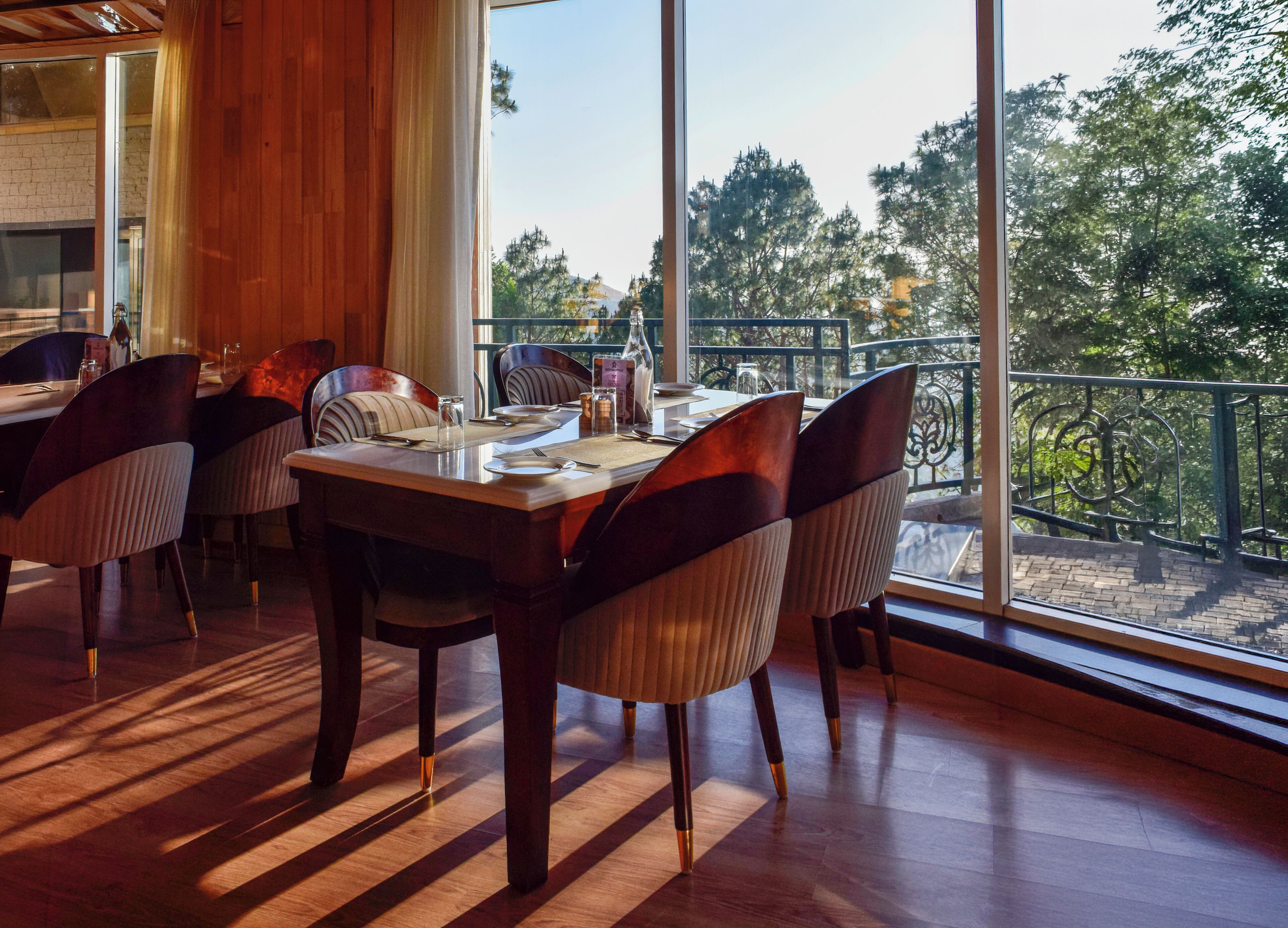 Un comedor con mesa y sillas de madera, ventanas amplias con vista a árboles y un edificio.
