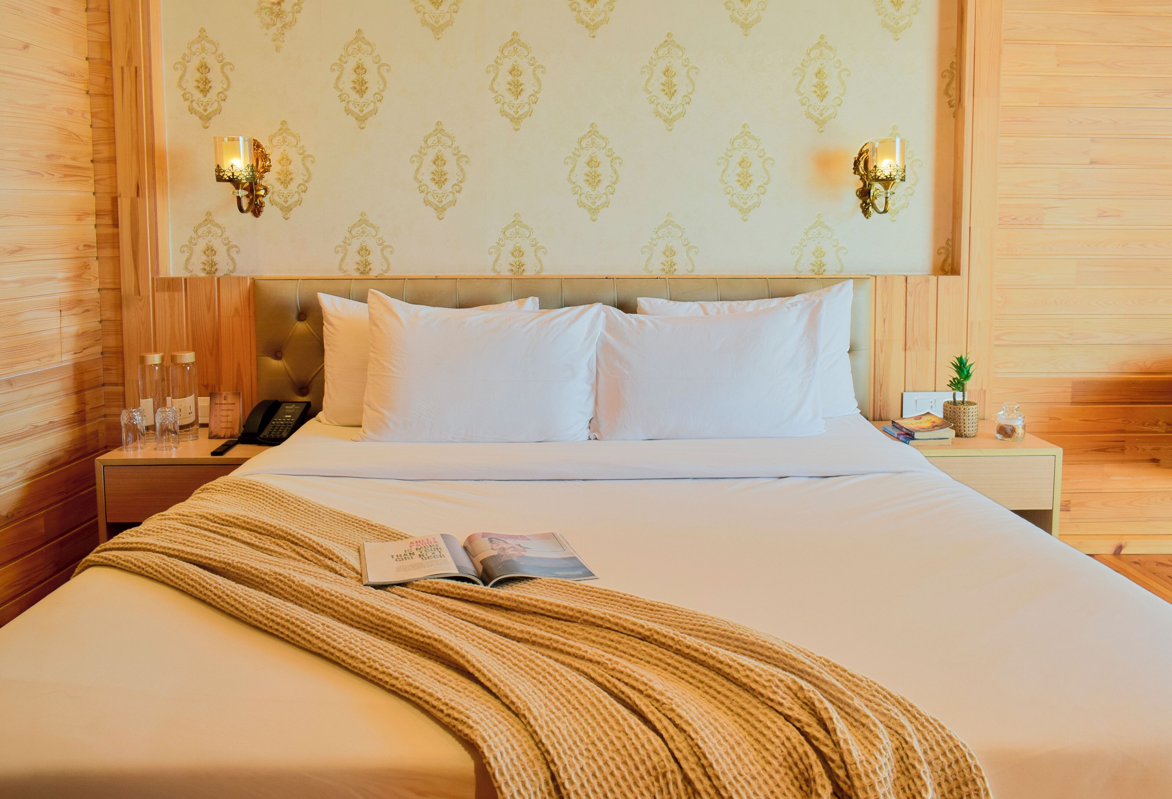 Quarto de hotel com cama, mesa de cabeceira com abajur, relógio de parede e uma pequena planta em vaso.