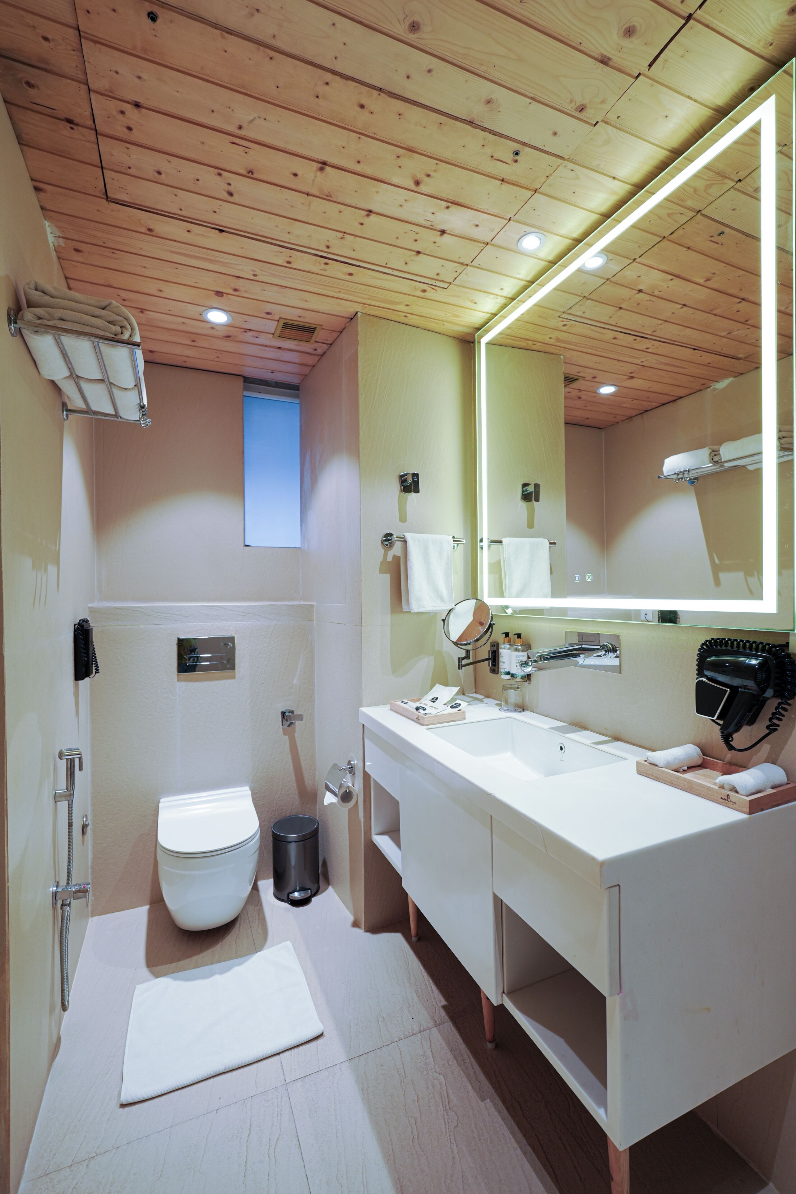 Un baño moderno con techo de madera, un espejo grande, un lavamanos blanco y un inodoro.