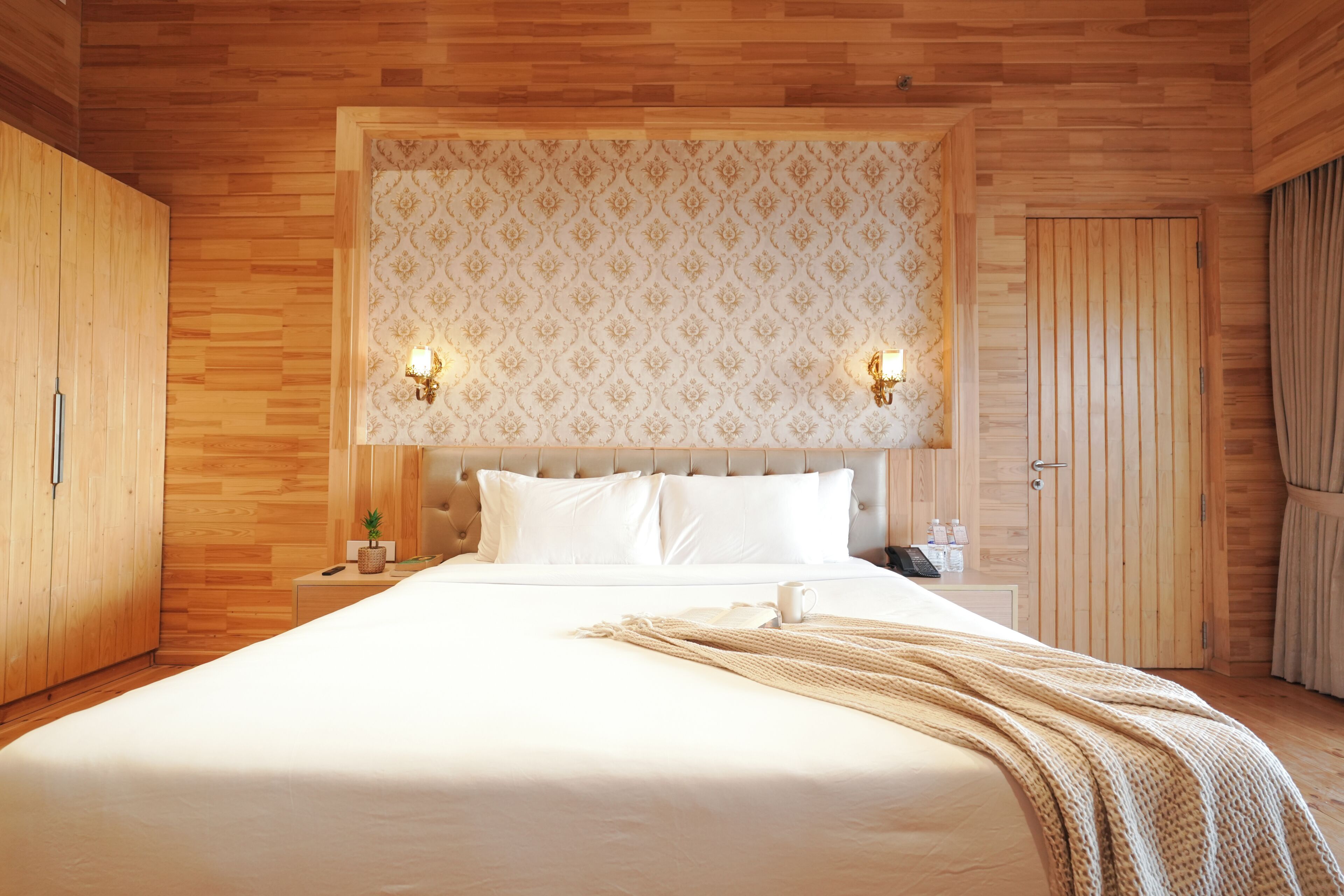 Habitación de hotel con techo de madera, una cama grande con ropa blanca, dos lámparas de pared, una mesita de noche con un teléfono y una planta pequeña, y un armario de madera.