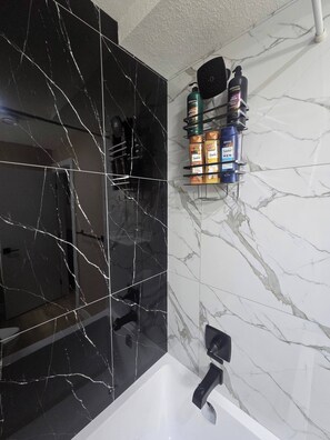 Une salle de bain avec un mur en marbre noir, un mur en marbre blanc et un lavabo noir. Plusieurs bouteilles sont disposées sur une étagère.