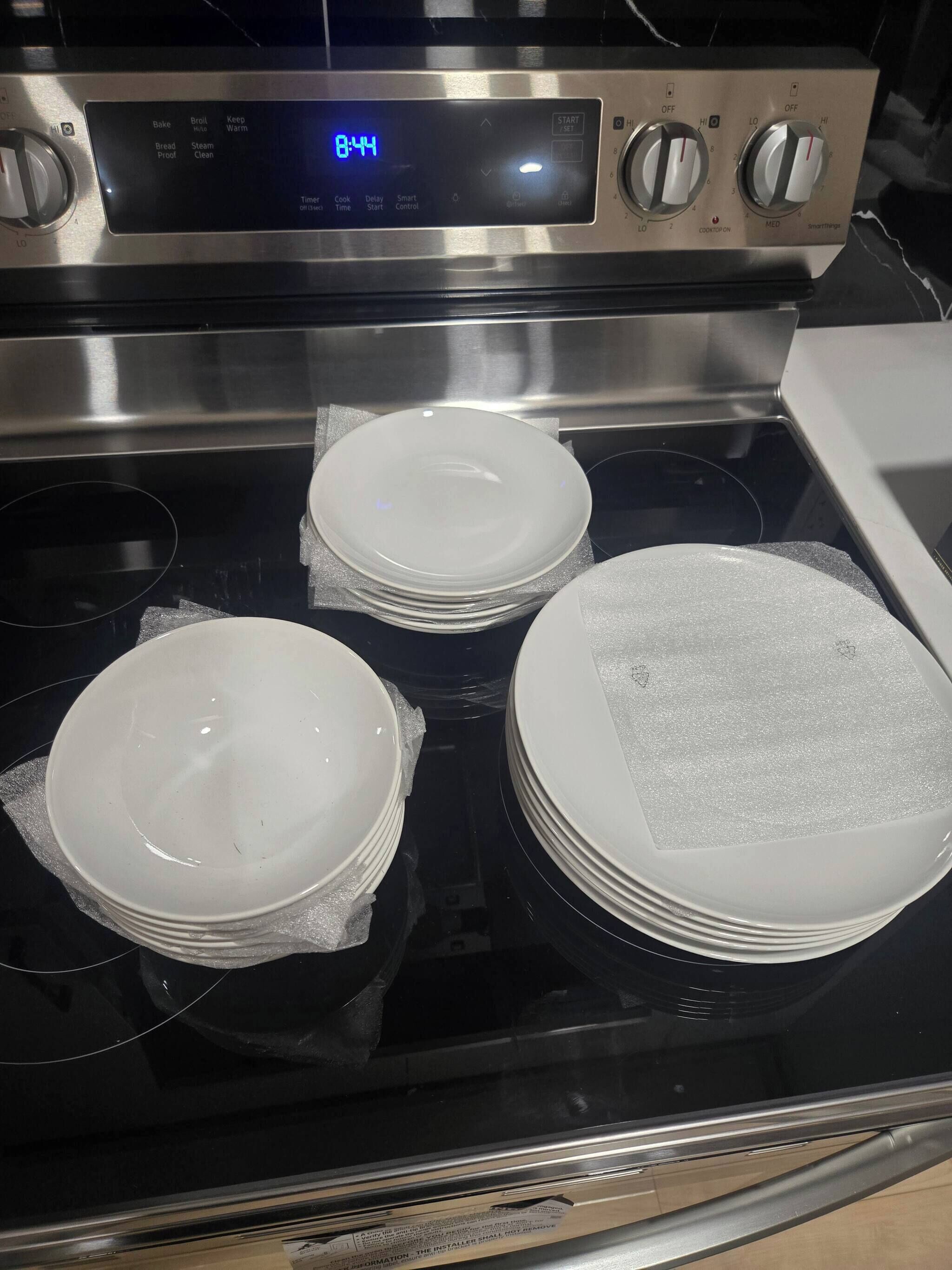 A stainless steel oven with a digital clock displaying 8:44, three stacked white plates on the oven's surface, and knobs for temperature and timer settings.