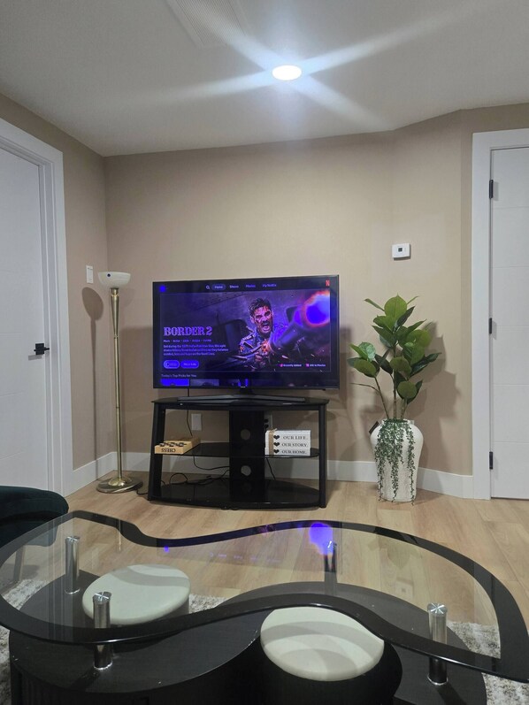 Un salon avec un téléviseur à écran plat posé sur un meuble, une table basse en verre, une plante en pot et un ventilateur de plafond.