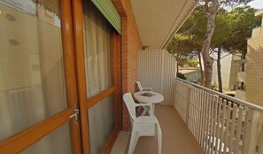 Ein Balkon mit Tisch und zwei Stühlen, eine Holztür und Blick auf Bäume und Gebäude.