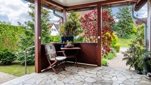 A covered patio with a stone floor, a wooden table, a chair, and a potted plant.