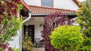 A house with a tiled roof, a front door, a small porch, and various potted plants.