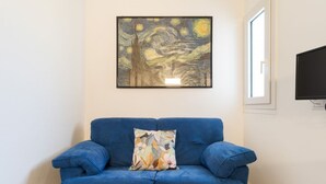 Een blauwe bank met een gebloemd kussen, een kunstwerk erboven en een televisie aan de muur.