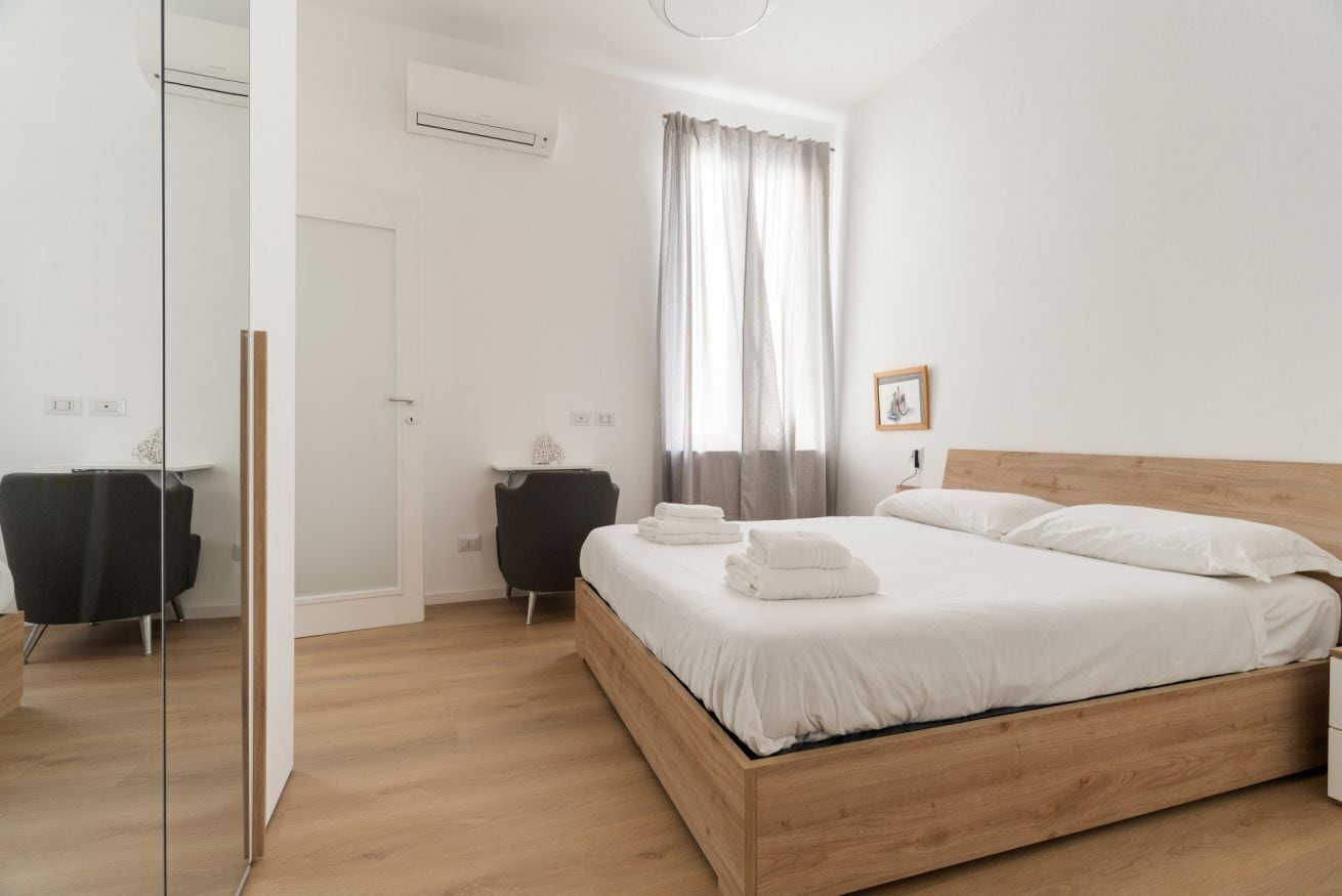 A modern bedroom with a wooden bed, white linens, a small bedside table, a chair, and a window with curtains.