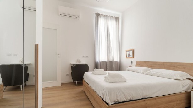 Een moderne slaapkamer met een houten bed, witte lakens, een klein nachtkastje, een stoel en een raam met gordijnen.