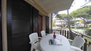 A round table with white chairs on a balcony, a closed dark wooden door, and a view of buildings and trees. - Spacious stay in a quiet area of Lignano Pineta (Lignano Sabbiadoro)