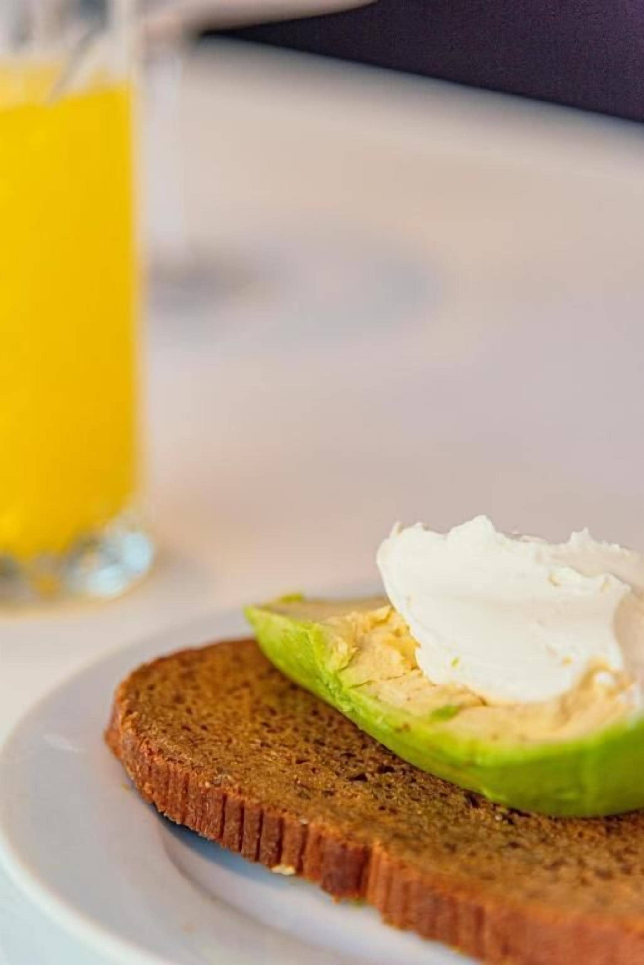 A slice of whole grain toast with avocado and a dollop of cream cheese on a white plate.