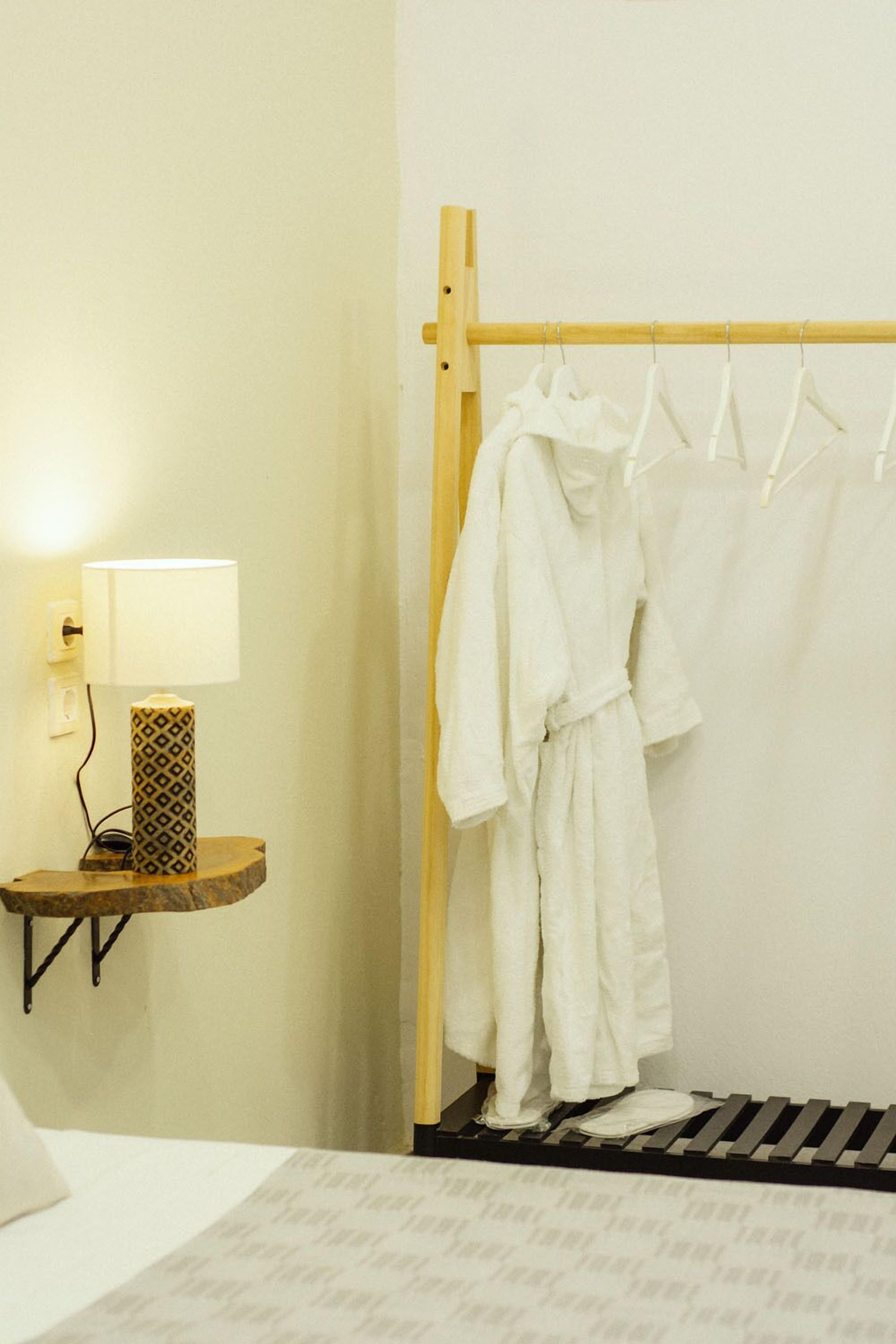 Ein Badezimmer mit einem hölzernen Ablagegestell für weiße Bademäntel, einem Nachttisch mit Lampe und einem Bett mit gemustertem Überwurf.