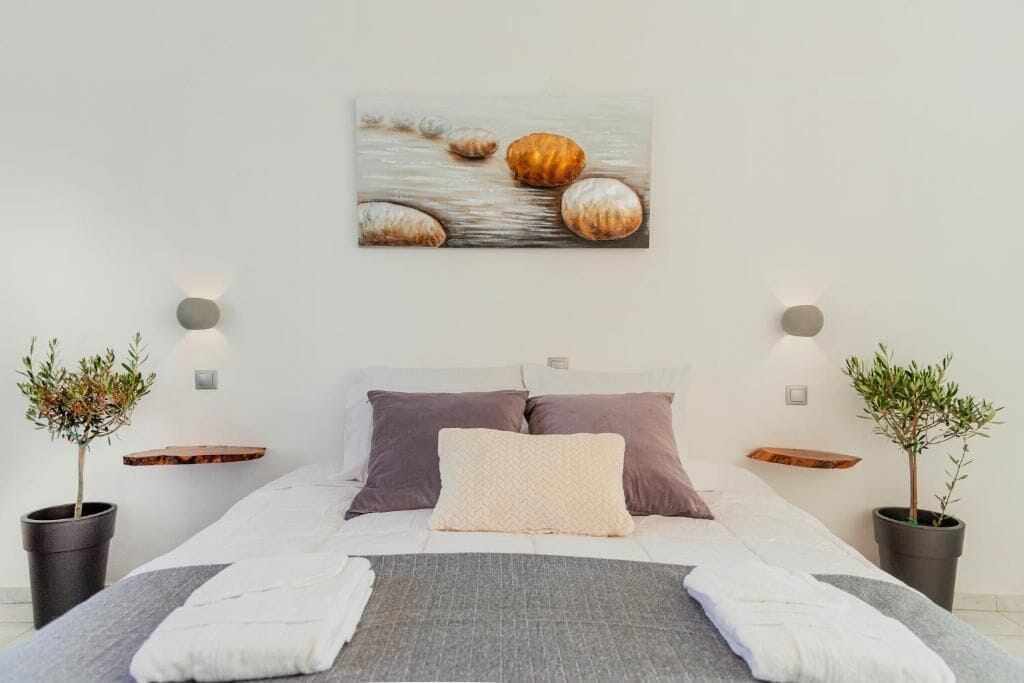 A bedroom with a bed, pillows, a blanket, bedside tables, wall-mounted lights, and a painting.