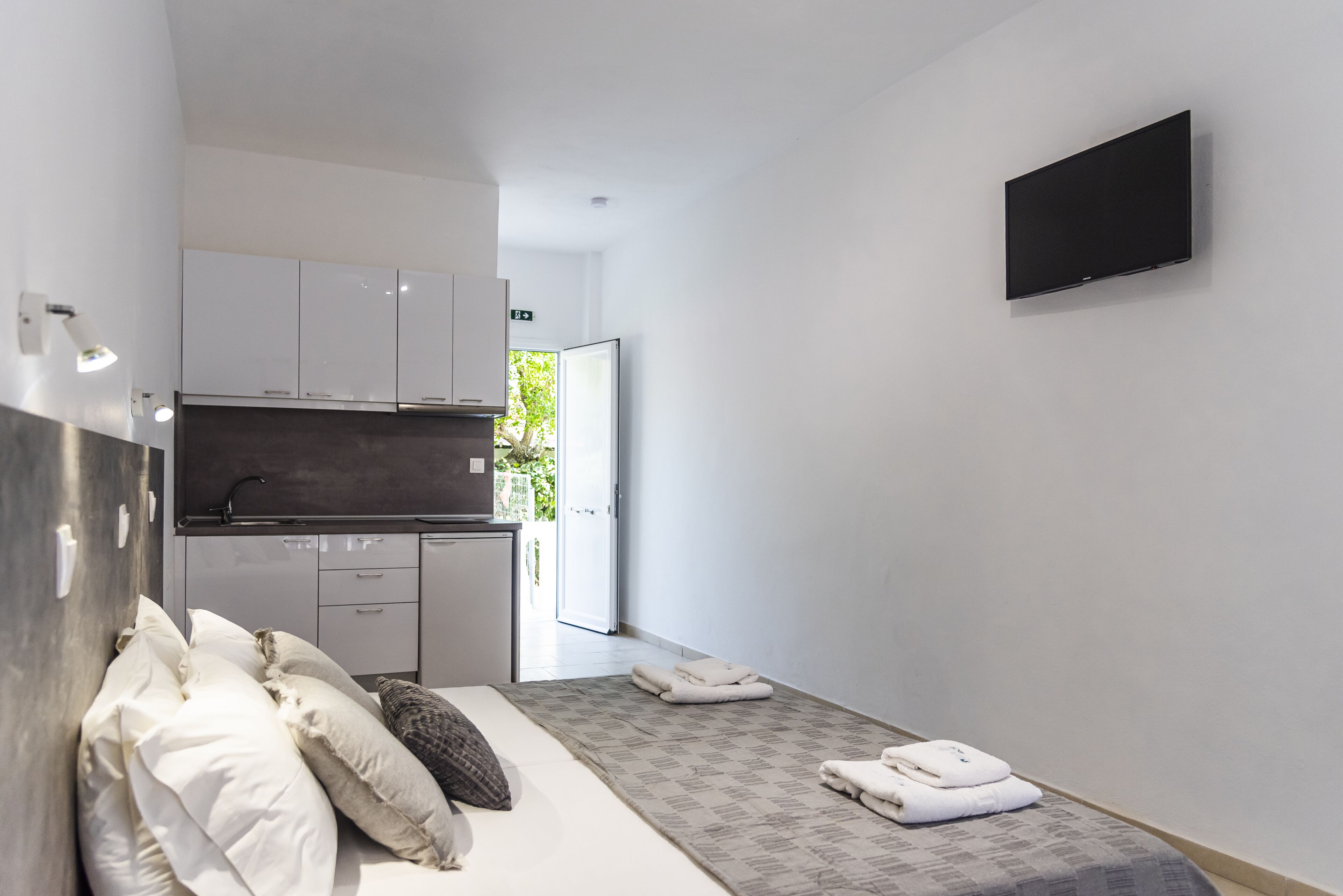 A modern hotel room with a bed, a flat-screen TV, a kitchenette, and a bathroom.