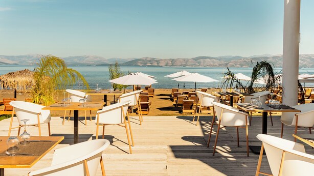 A seaside restaurant with outdoor seating, tables, chairs, umbrellas, and a view of the sea and mountains. - Ionian Blue Beach - Adults Only (Corfu)