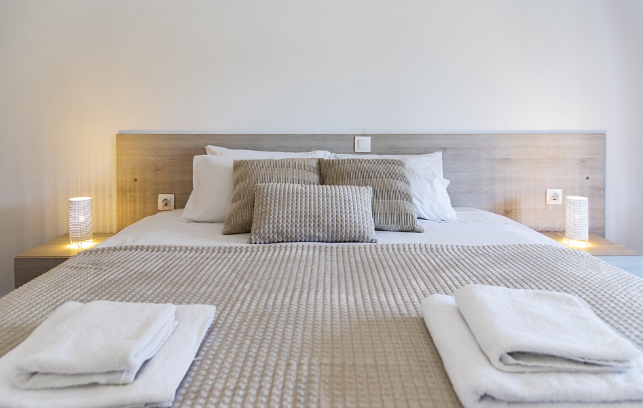 A neatly made bed with a patterned bedspread, two pillows, and two folded towels on a wooden headboard.
