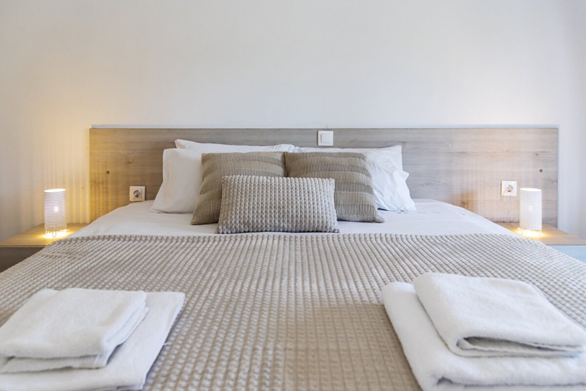 Una cama bien hecha con un edredón estampado, dos almohadas y dos toallas dobladas sobre un cabecero de madera.