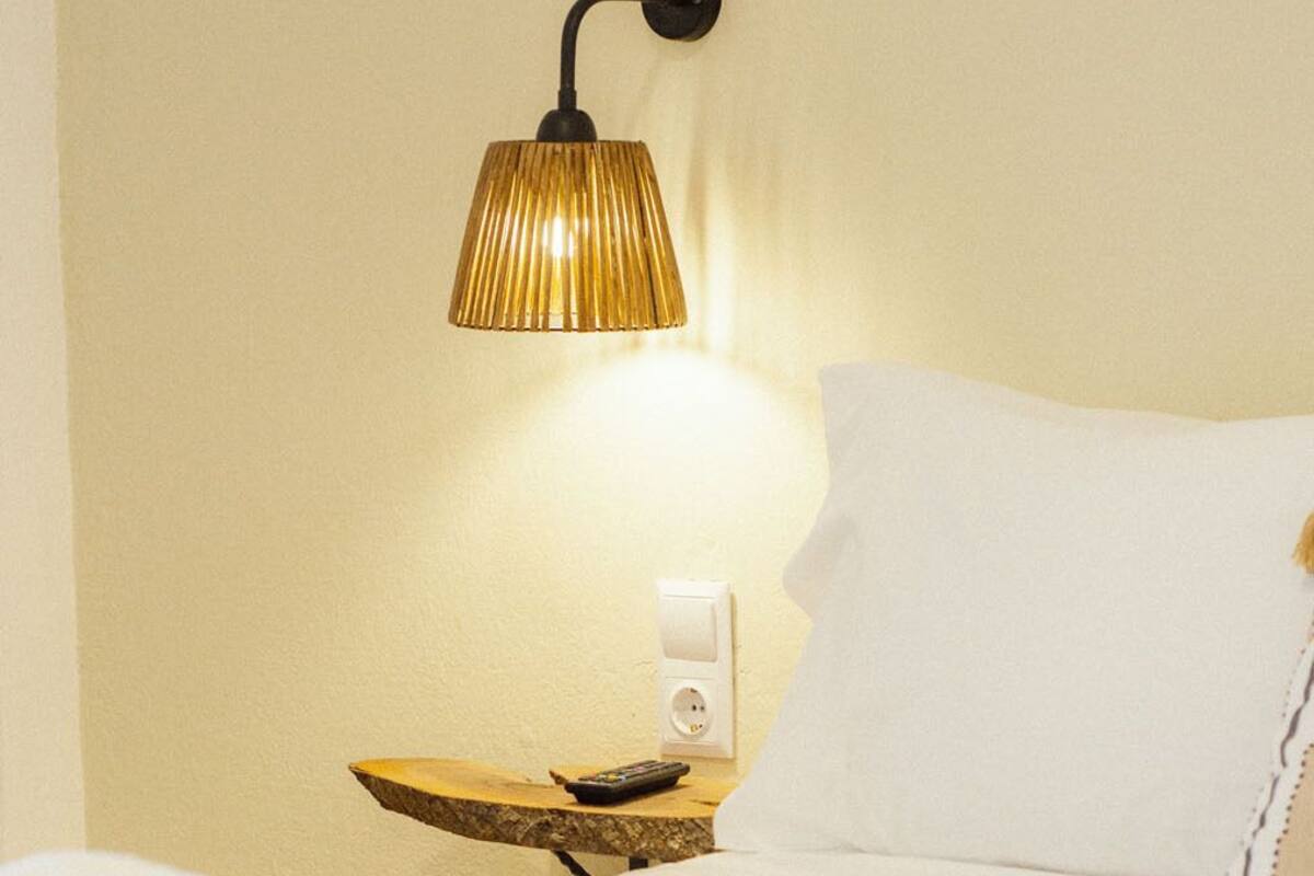 A hotel room with a bed, a wall-mounted lamp, a remote control, and a power outlet.
