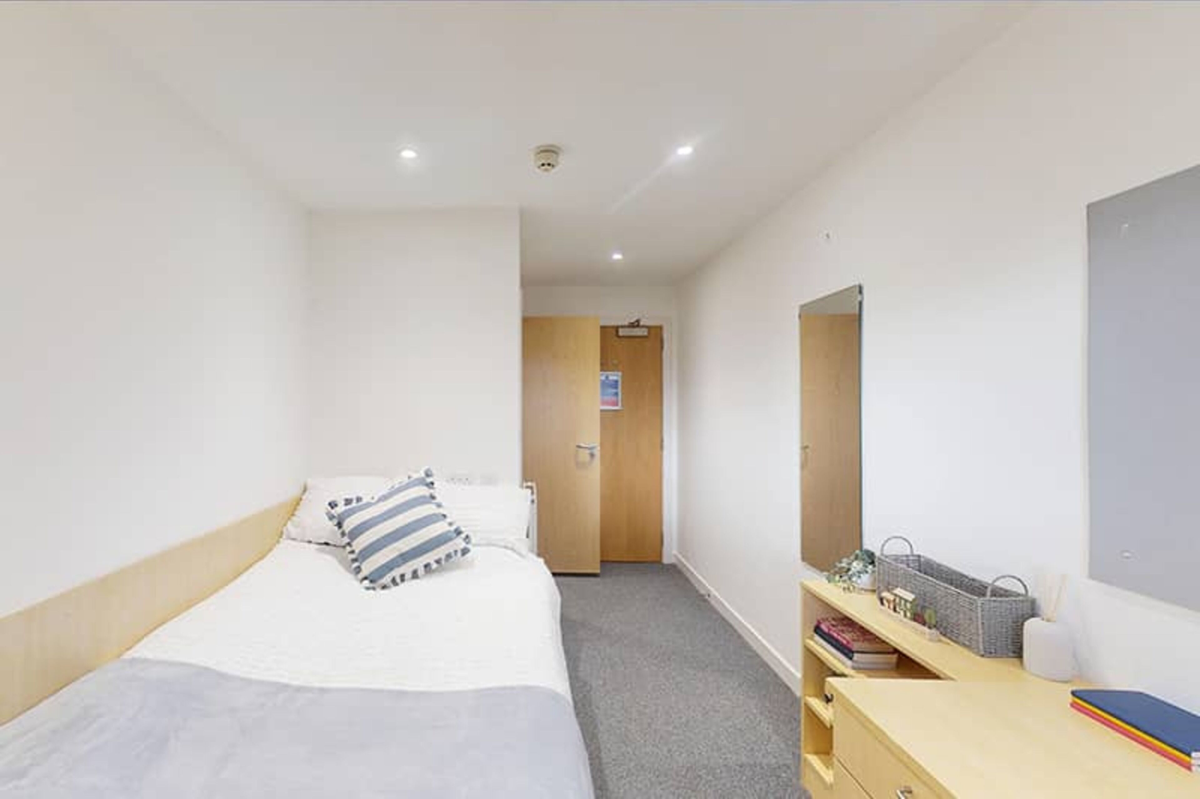 A hotel room with a bed, a desk with a basket, a mirror, and a door leading to a corridor.