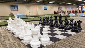 Ein großes Schachbrett im Innenbereich mit Figuren, eine rote Stange und im Hintergrund Spielautomaten.