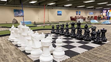 Een groot binnenshuis schaakbord met stukken, een rode paal en speelautomaten op de achtergrond.