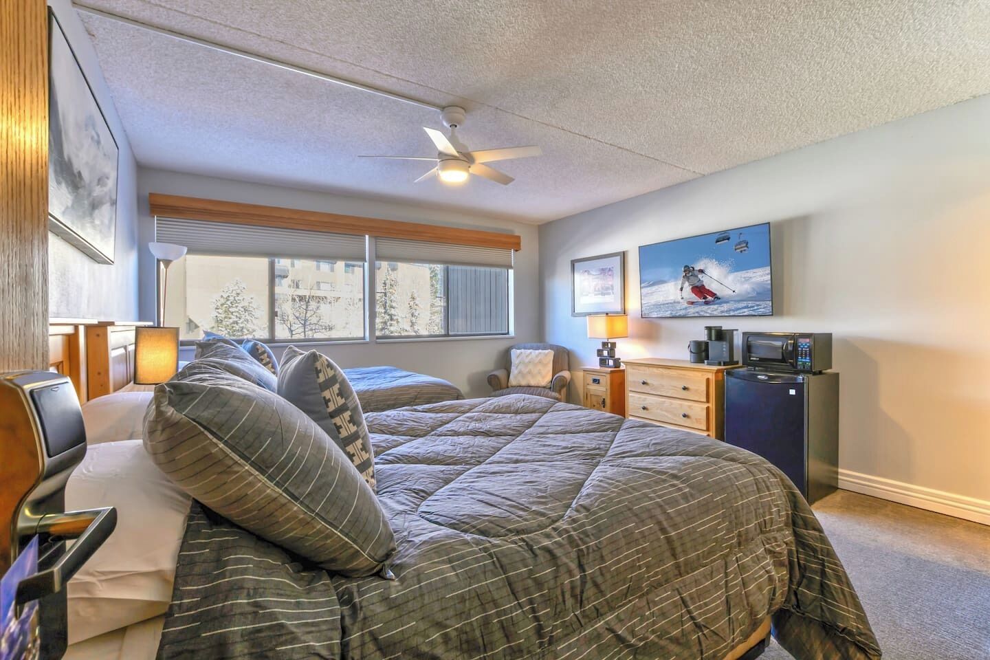Een slaapkamer met twee bedden, een plafondventilator, een raam met jaloezieën, een bureau met lamp, een dressoir, een magnetron en een ingelijst schilderij aan de muur.