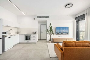 A modern kitchen with white cabinets, a microwave, and a built-in oven. There is a living area with a brown sofa and a large window. - Premium 2BR | Steps to Beach • Balcony • Mamad (Bat Yam)