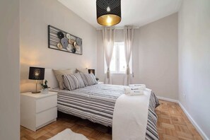 Ein Schlafzimmer mit einem Bett, Nachttischen, einer Lampe, einem Wandbild und einem Fenster mit Vorhängen.