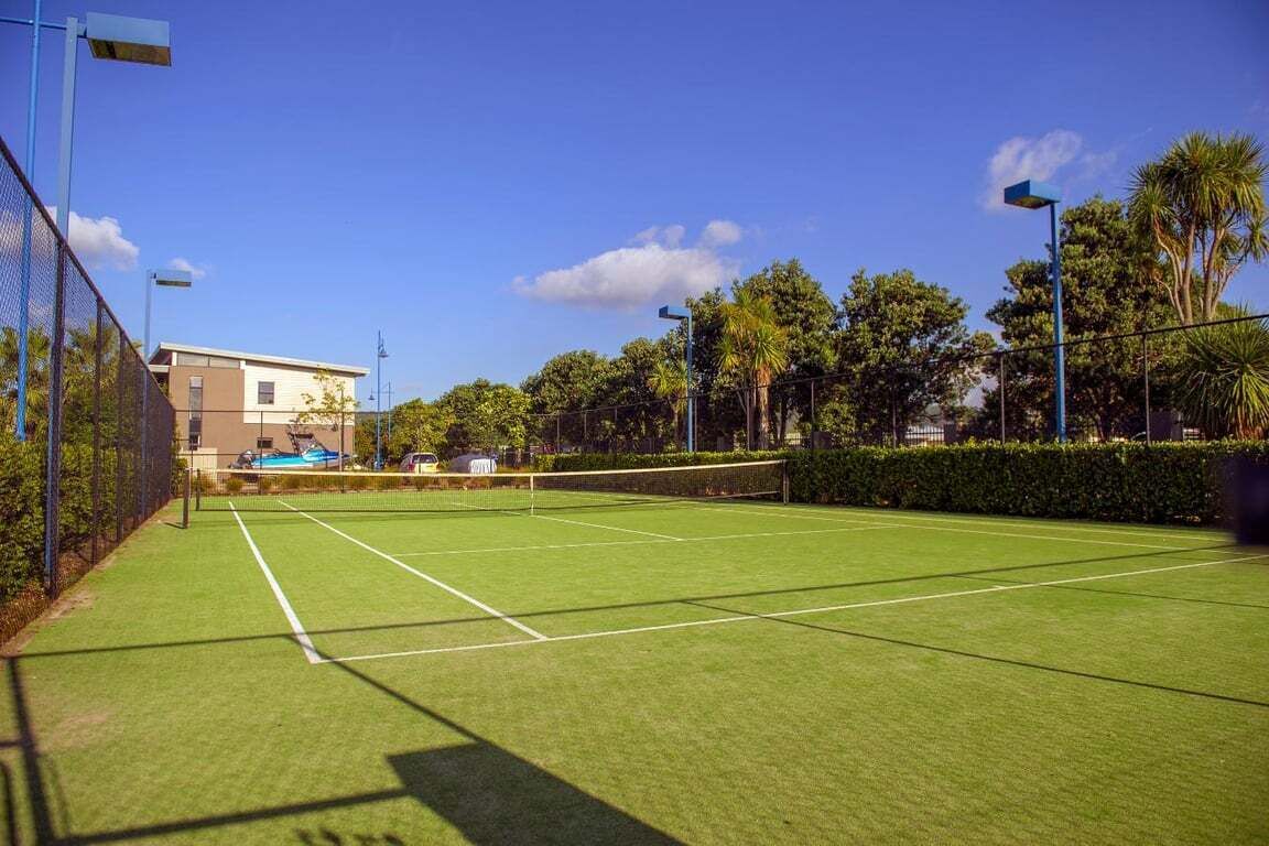 Una cancha de tenis con una red, rodeada por una cerca, árboles y un edificio al fondo.