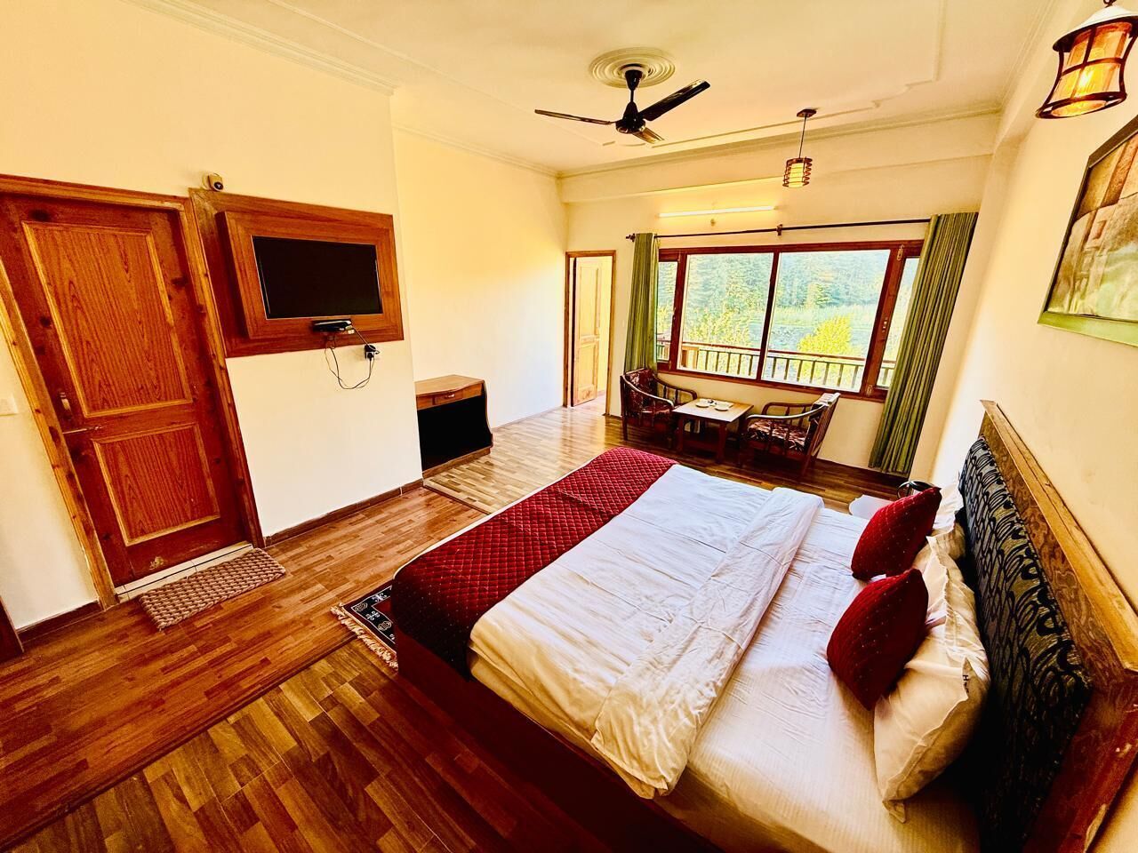 Un dormitorio con cama, piso de madera, ventilador de techo, televisión, balcón con vistas y una puerta.