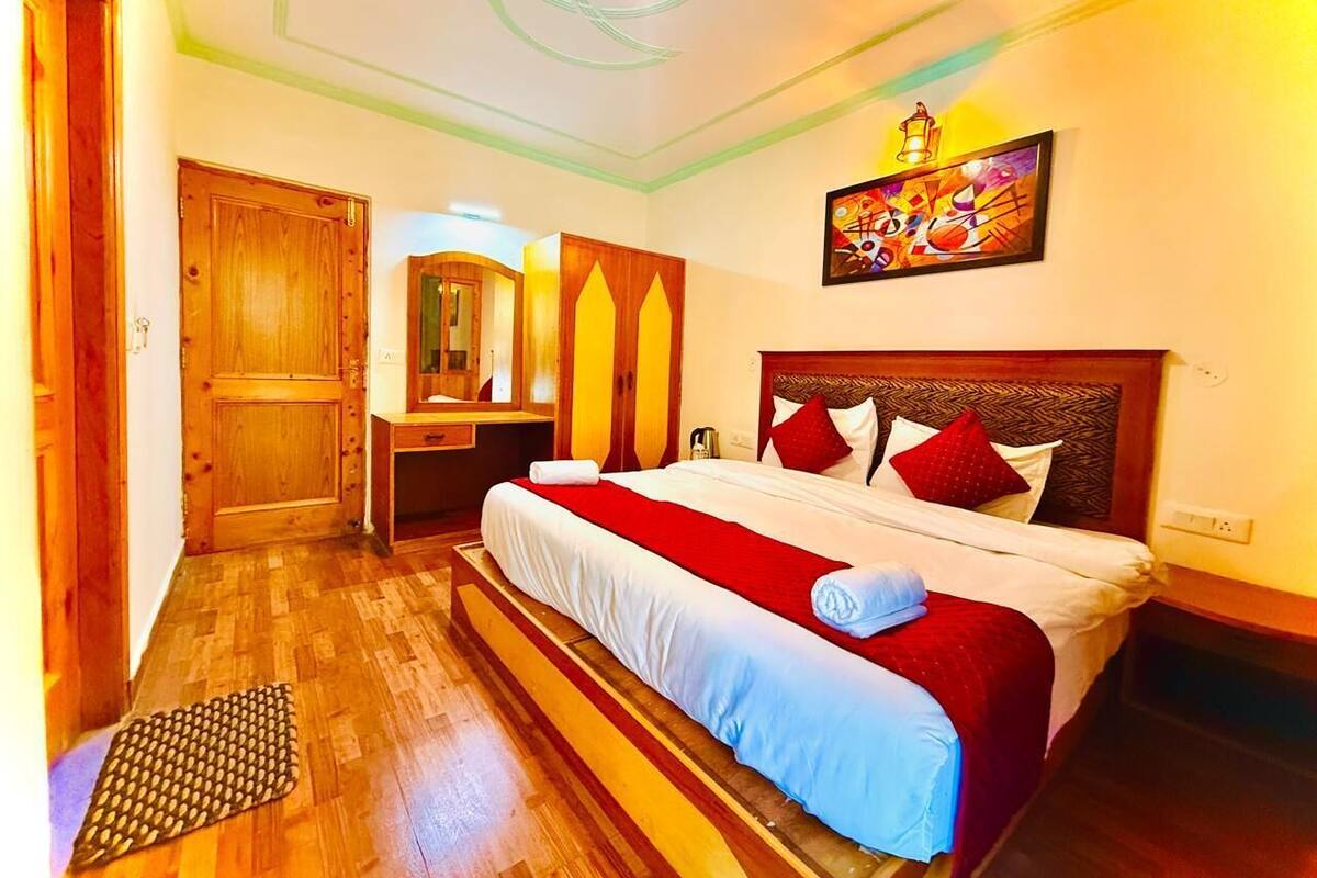 A hotel room with a wooden floor, a bed with red and white bedding, a wooden headboard, a bedside table, a mirror, and a painting on the wall.