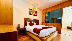 A wooden-framed bed with white bedding and red accents, a nightstand with a kettle, and a large window with a view. - The Manrals Resort (Manali)
