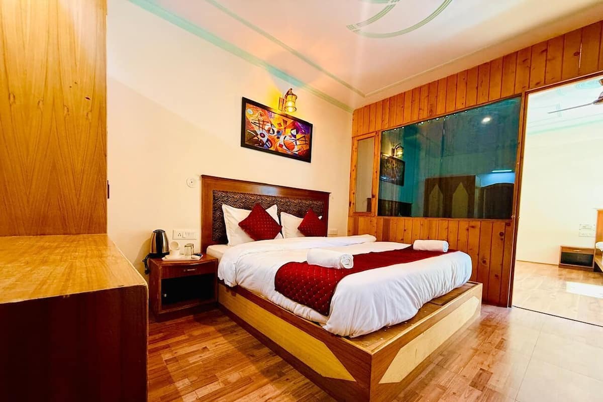 A wooden-framed bed with white bedding and red accents, a nightstand with a kettle, and a large window with a view.
