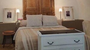 Un dormitorio con un armario de madera, una cama con colcha a rayas, un aparador blanco con cajones y dos mesitas de noche con lámparas.