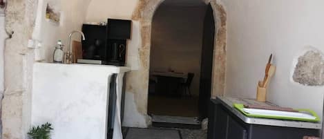 En smal, välvd dörröppning som leder till en matplats, ett litet kök med en diskho och en arbetsbänk samt ett svart skåp med ett grönt bricka.