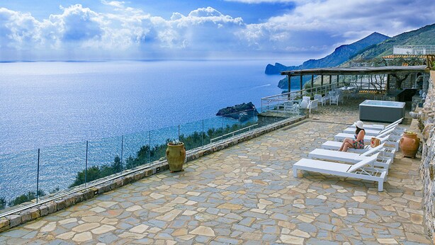 Une terrasse dallée de pierres, agrémentée de chaises longues blanches, offre une vue imprenable sur la mer, une côte rocheuse et un paysage montagneux en arrière-plan.