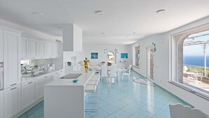 Une cuisine moderne avec des meubles blancs, un îlot central et une vue sur la mer grâce à de grandes fenêtres.