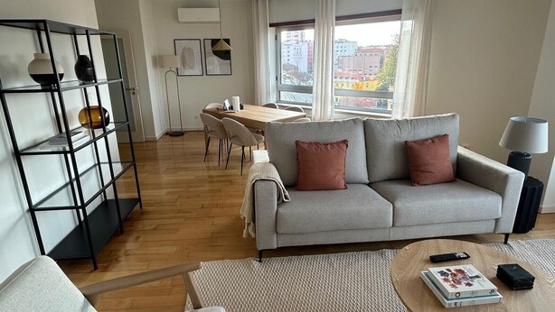 Een moderne woonkamer met een grijze bank, een salontafel, een boekenkast en een eethoek met een houten tafel en stoelen.
