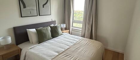 A bedroom with a bed, two bedside tables, a window with curtains, and two framed abstract artworks on the wall.