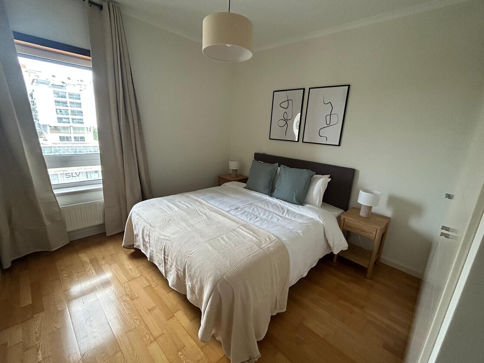 Un dormitorio con una cama grande, dos mesitas de noche, una ventana con cortinas y dos cuadros decorativos.